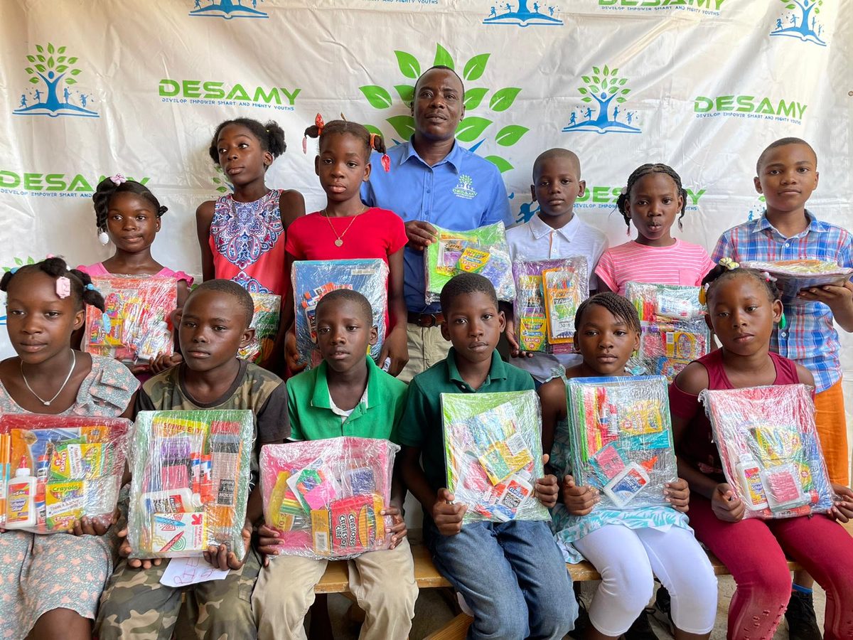 Children holding school supply kits with a DESAMY team member in front of a DESAMY banner in Haiti